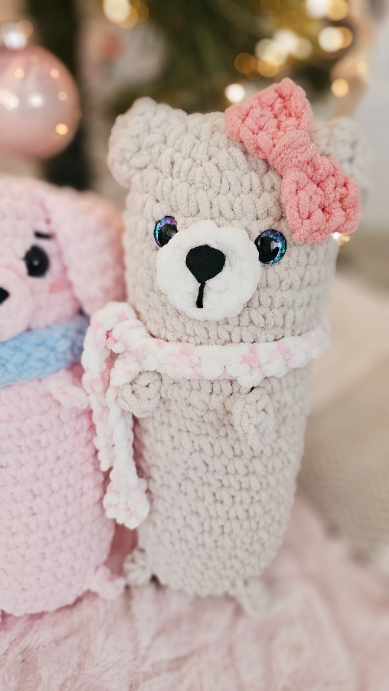 May include: A crocheted gray bear with a pink bow and a pink and white scarf. The bear is sitting on a pink fuzzy surface.