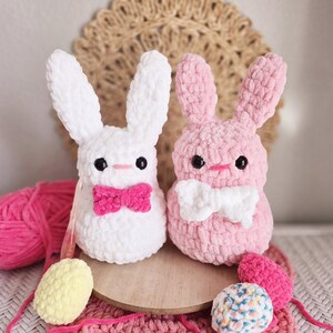 May include: Two crocheted bunny plushies, one white with a pink bow and one pink with a white bow. The plushies are sitting on a wooden surface with yarn balls nearby.