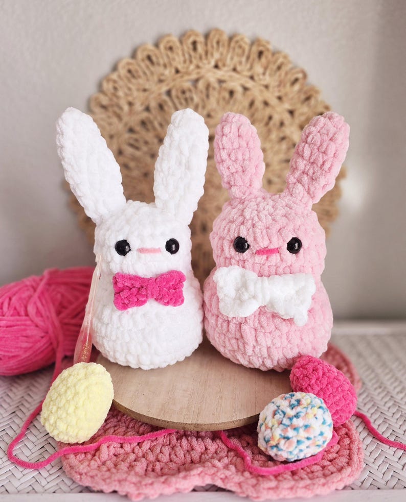 May include: Two crocheted bunny plushies, one white with a pink bow and one pink with a white bow. The plushies are sitting on a wooden surface with yarn balls nearby.