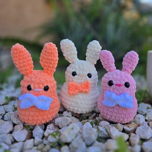 May include: Three crocheted bunny plushies with different colored fur. The bunnies are wearing blue bow ties. The bunnies are sitting on a bed of small rocks.