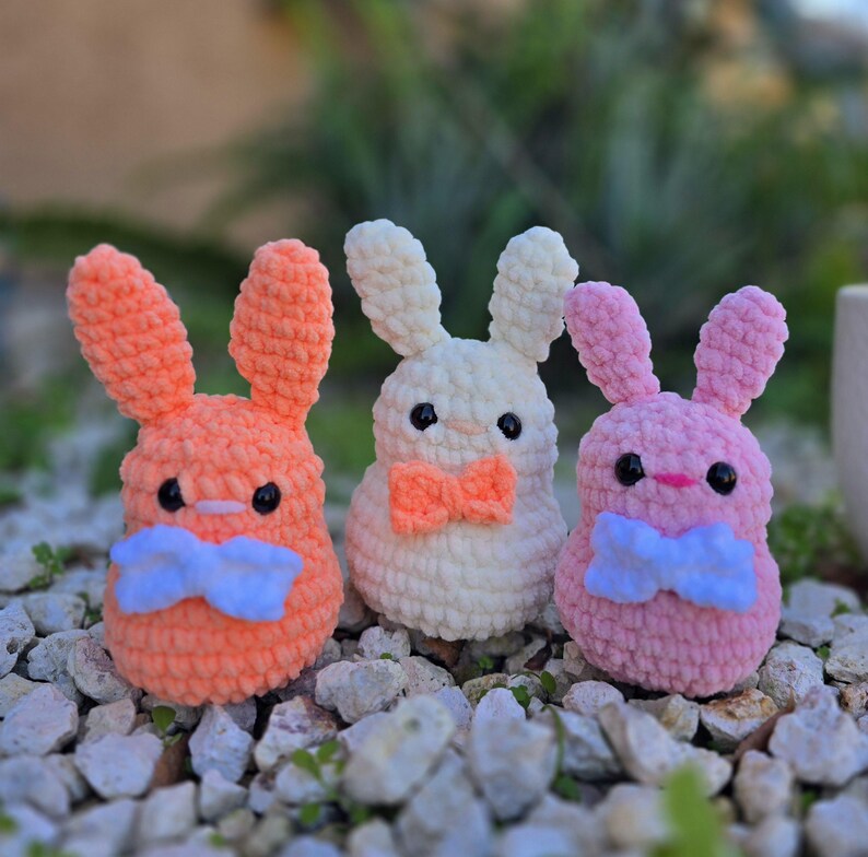 May include: Three crocheted bunny plushies with different colored fur. The bunnies are wearing blue bow ties. The bunnies are sitting on a bed of small rocks.