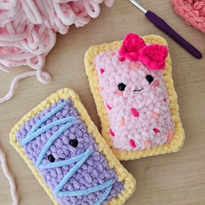 Puede incluir: Dos juguetes de peluche tejidos a crochet con forma de tartas. Uno es rosa con un lazo rosa y el otro es morado con glaseado azul. Ambos tienen una corteza amarilla y caras sonrientes.