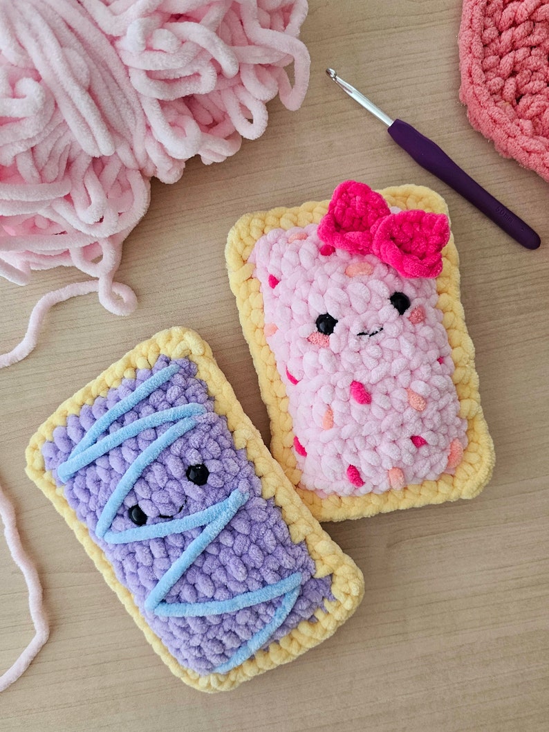 Puede incluir: Dos juguetes de peluche tejidos a crochet con forma de tartas. Uno es rosa con un lazo rosa y el otro es morado con glaseado azul. Ambos tienen una corteza amarilla y caras sonrientes.