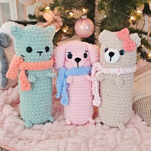 May include: Three crocheted stuffed animals: a light blue cat, a pink dog, and a beige bear. Each animal is wearing a scarf and has embroidered eyes. The animals are sitting on a fluffy pink surface.