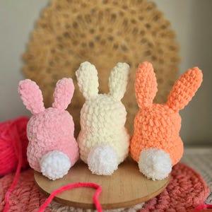 May include: Three crocheted bunny rabbits in pink, white, and orange. Each bunny has a white pom-pom tail.
