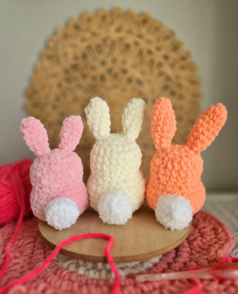 May include: Three crocheted bunny rabbits in pink, white, and orange. Each bunny has a white pom-pom tail.