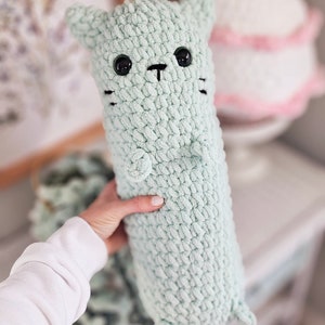 May include: A soft, crocheted green cat toy with black eyes and a black nose. The cat is being held by a hand.