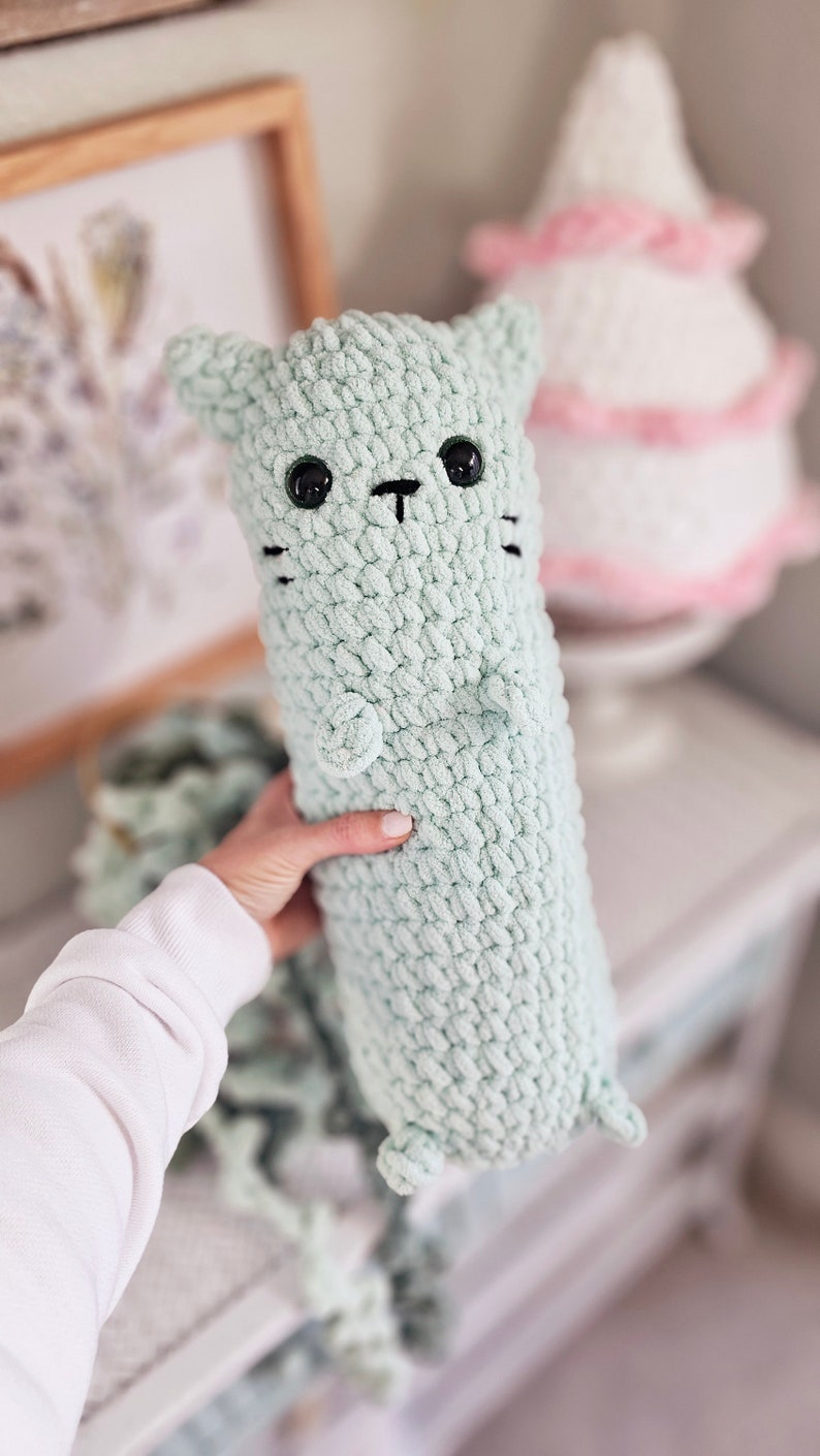 May include: A soft, crocheted green cat toy with black eyes and a black nose. The cat is being held by a hand.