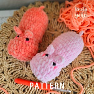 May include: Two crocheted plush hippopotamus toys, one pink and one orange, on a woven mat with a crochet hook and yarn. The text "knots & yarn" is visible in the upper right corner of the image. The text "PATTERN" is visible in the lower center of the image.