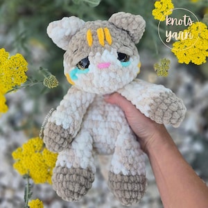 Può includere: Un giocattolo a forma di gatto di peluche fatto a mano con un'espressione triste. Il gatto è beige, bianco e marrone con strisce gialle sulla testa e accenti blu e rosa. Il giocattolo è tenuto in mano, con fiori gialli sullo sfondo. Il testo "knots yanni" è in alto a destra.
