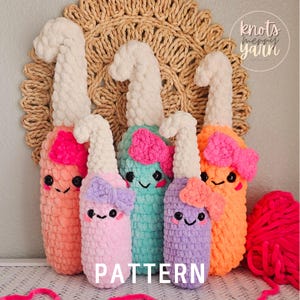 Puede incluir: Cinco juguetes de peluche tejidos a crochet con forma de botellas, con hilo de diferentes colores y caras lindas. Los juguetes están dispuestos en fila sobre una estera tejida con el texto "knots merry yarn" en el fondo.