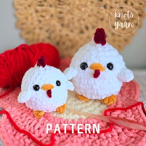 May include: Two white crocheted chicken amigurumi toys with red combs and orange beaks. The smaller chicken is sitting on a pink and white crocheted blanket with a crochet hook. The larger chicken is standing on the blanket. The text "PATTERN" is printed on the blanket.