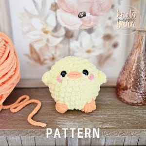 May include: A crocheted yellow chick amigurumi toy with pink cheeks and a small orange beak. The chick is sitting on a wooden surface with a ball of yarn and a glass vase in the background. The text "PATTERN" is written below the chick.