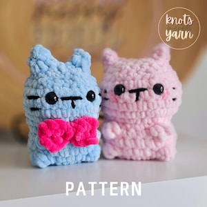 May include: Two crocheted cat plushies, one blue with a pink bow tie and one pink. The plushies have black eyes and a black nose. The image includes the text "knots merry yarn".