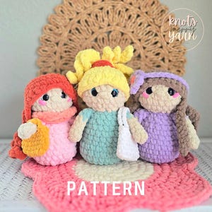 May include: Three handmade crochet dolls in pink, blue, and purple, each with unique hats and accessories. The dolls are on a pink and white crocheted mat with the word "PATTERN" in white. The background includes a brown woven placemat and the text "knots merry yarn".