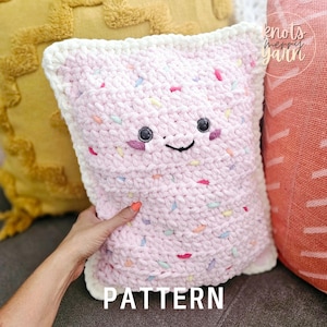 May include: A pink crocheted pillow shaped like a square with a smiling face and colorful sprinkles. The pillow has a white border and the text "PATTERN" is printed below it.