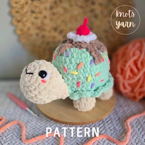 May include: A crocheted turtle toy with a green shell that looks like ice cream with sprinkles. The turtle has a beige head and a cherry on top of the ice cream.