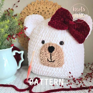 Op de afbeelding: Witte gehaakte beren knuffel met een bruine neus en ogen. De beer draagt een grote bordeauxrode strik en de tekst "knots merry yarn" is zichtbaar in de rechterbovenhoek van de afbeelding. De tekst "PATTERN" is zichtbaar in het midden onderaan de afbeelding.