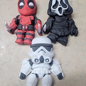 Peut inclure: Trois petites figurines : Deadpool en rouge avec des détails noirs et deux couteaux argentés, Ghostface en noir et blanc, et un Stormtrooper en blanc et noir. Les figurines sont stylisées et ont un aspect de dessin animé.