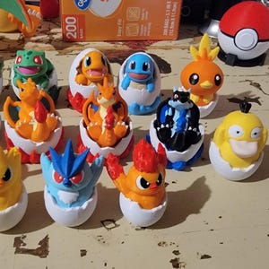 Może przedstawiać: Kolekcja 12 figurek Pokemonów w pojemnikach w kształcie jajka. Figurki są w różnych kolorach, w tym czerwonym, pomarańczowym, żółtym, zielonym, niebieskim i fioletowym. Każda figurka jest w innej pozie, a niektóre mają zamknięte oczy. Figurki znajdują się w białym pojemniku w kształcie jajka z białą podstawą.