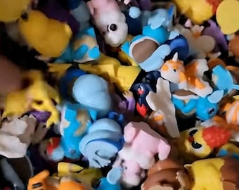 May include: A collection of colorful, small figurines in various shapes and designs. The figurines are primarily blue, yellow, pink, and brown, with some featuring horns, wings, and other unique features. The image is a close-up, showing the figurines densely packed together.