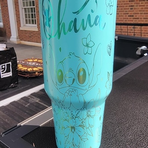 Puede incluir: Un vaso de color turquesa con un diseño del personaje Stitch de Lilo & Stitch, con la palabra "Ohana". El diseño incluye detalles florales y de corazón. El vaso tiene una tapa blanca y una banda turquesa metálica.