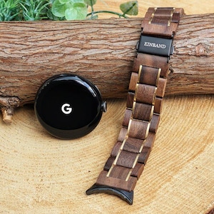 May include: A dark brown wooden watch band with gold accents and a black metal buckle. The band has the word "EINBAND" printed on the buckle. The band is lying on a wooden surface.