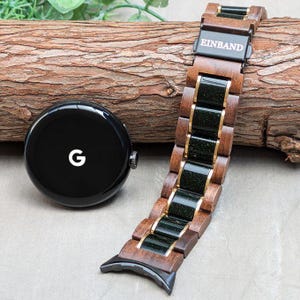 May include: A dark brown wooden watch band with a black and green inlay. The band has gold accents and the text "EINBAND" is printed on the band. The band is shown next to a black Google smartwatch.