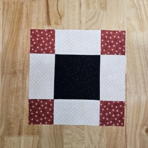 Może przedstawiać: Kwadratowy blok do pikowania z czarnym środkiem, białą tkaniną w kropki i czerwonymi narożnikami w kropki. Blok składa się z zszytych ze sobą kwadratów materiału, prezentujących prosty, ale klasyczny wzór. Blok jest prawdopodobnie częścią większego projektu pikowania.