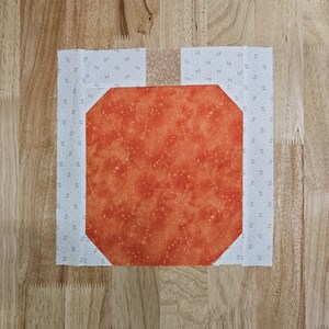 May include: A square quilt block featuring a central orange octagonal shape with a mottled pattern. The orange shape is surrounded by a white fabric with a small, repeating gray pattern. A tan strip is at the top and bottom.