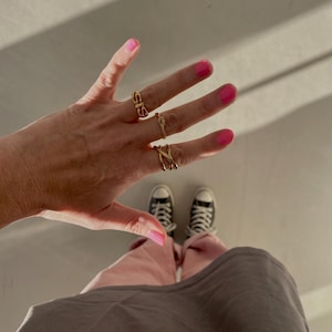 Könnte beinhalten: Eine Hand trägt vier goldene Ringe, einer davon mit dem Wort "SIS" darauf. Die Nägel sind in einem leuchtenden Pink lackiert.