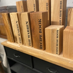 May include: A wooden shelf unit with two drawers and multiple shelves. The shelves are filled with wooden blocks with various text labels, including "The Warren", "Recipe Book", and "Johns Music Room".