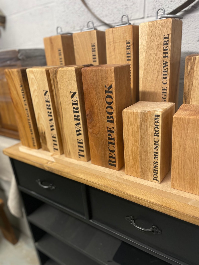 May include: A wooden shelf unit with two drawers and multiple shelves. The shelves are filled with wooden blocks with various text labels, including "The Warren", "Recipe Book", and "Johns Music Room".