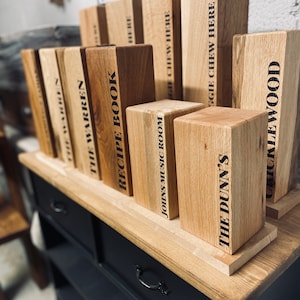 May include: A collection of wooden blocks with various text labels, including "The Warren", "Recipe Book", "Johns Music Room", "The Dunn's", and "Chucklewood". The blocks are arranged on a wooden shelf with a black painted surface.