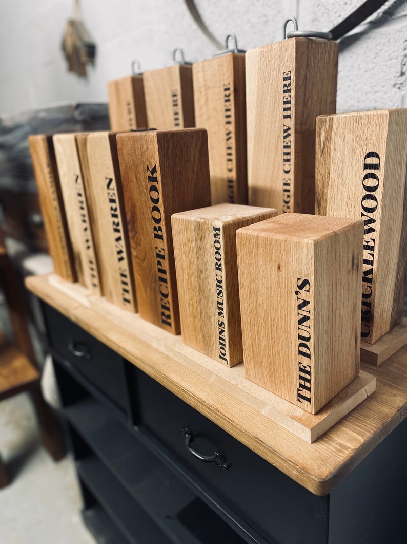 May include: A collection of wooden blocks with various text labels, including "The Warren", "Recipe Book", "Johns Music Room", "The Dunn's", and "Chucklewood". The blocks are arranged on a wooden shelf with a black painted surface.