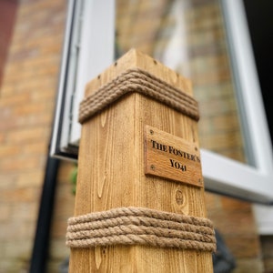 May include: A wooden mailbox, rectangular in shape, made from light brown wood. It features thick rope detailing around the top and base. A small wooden plaque displays the text "The Foster's YO41".