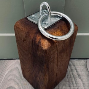May include: A dark brown wooden doorstop with a silver metal ring attached to the top. The ring is attached to a square metal plate with screws.