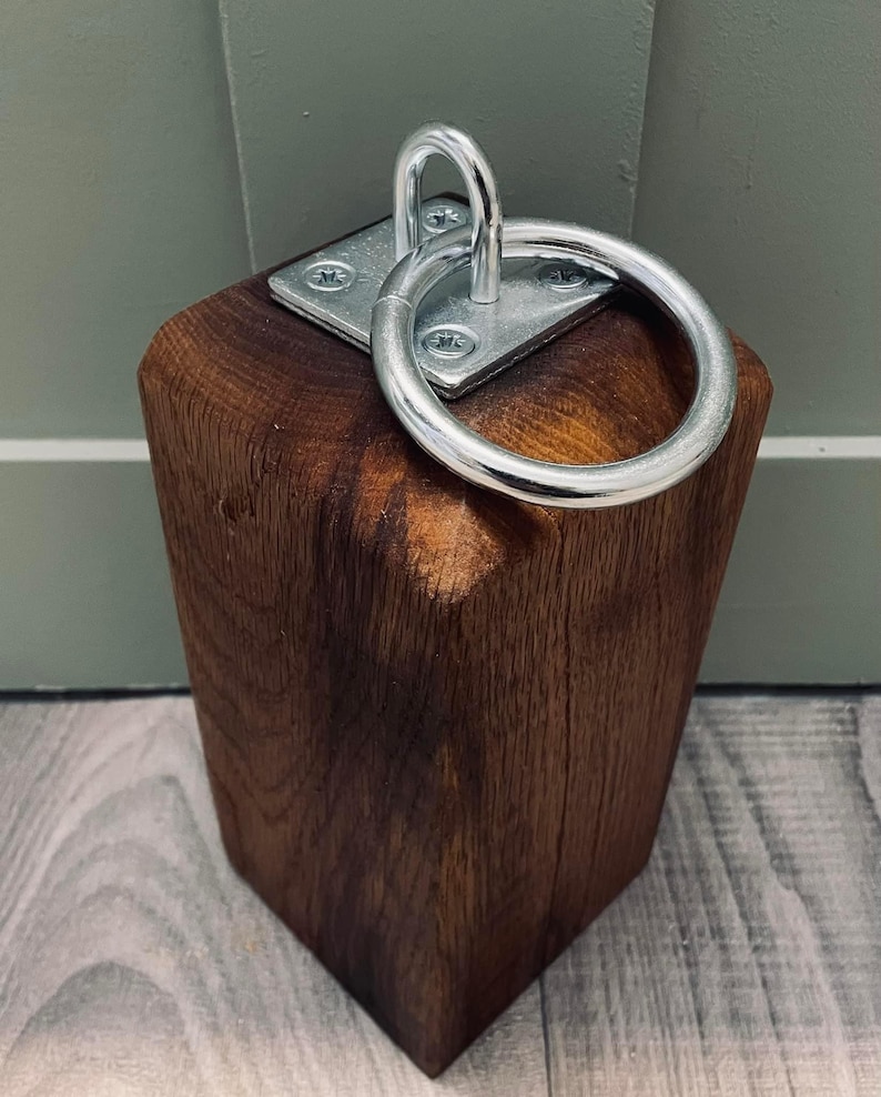 May include: A dark brown wooden doorstop with a silver metal ring attached to the top. The ring is attached to a square metal plate with screws.