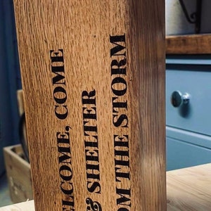 May include: A wooden doorstop with a metal ring on top. The doorstop is engraved with the text "Welcome, Come In & Shelter From The Storm".