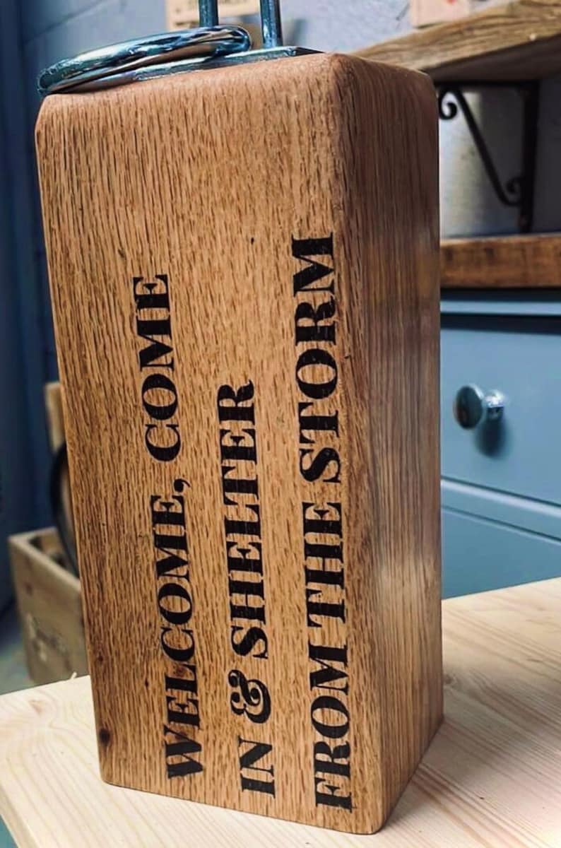 May include: A wooden doorstop with a metal ring on top. The doorstop is engraved with the text "Welcome, Come In & Shelter From The Storm".