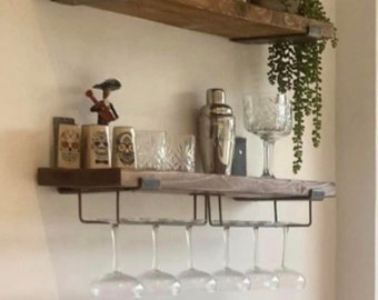 May include: A wooden shelf with a metal wine glass rack, holding six wine glasses. The shelf is decorated with bottles of spirits, a cocktail shaker, and a small decorative figurine. The shelf is mounted on a white wall.