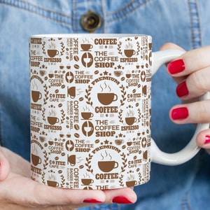 May include: White ceramic coffee mug with a brown and white design featuring the text "The Coffee Shop" and other coffee-related words and graphics.