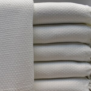 May include: A stack of white woven blankets with fringed edges. The blankets are soft and textured, and they would be perfect for a cozy night in.