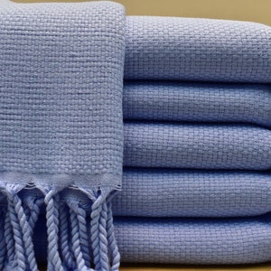 May include: A stack of light blue woven towels with fringed edges. The towels have a textured appearance and are neatly folded, showcasing a clean and organized presentation. The fringe adds a decorative element.