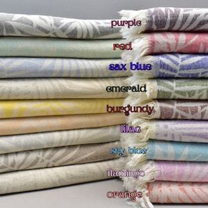 Puede incluir: Una pila de 12 toallas turcas en varios colores, incluyendo morado, rojo, azul saxo, esmeralda, burdeos, lila, azul cielo, flamenco y naranja. Cada toalla presenta un patrón de hojas blancas.