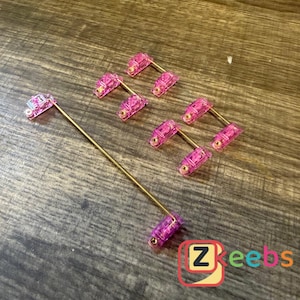 May include: A collection of pink and gold-colored accessories. The accessories have a rectangular pink top with a gold-colored stem. The accessories are arranged on a wooden surface. The logo "Zkeebs" is in the bottom right corner.