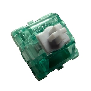 以下が含まれることがあります： 白いステムを持つ緑色のメカニカルキーボードスイッチ。スイッチは透明で、側面に「GATERON」という文字が印刷されています。