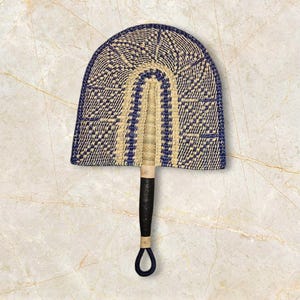 May include: A hand fan with a woven design in navy blue and beige. The fan has a black handle with a loop at the end. The fan's shape is a half-circle, and the woven pattern is visible throughout.