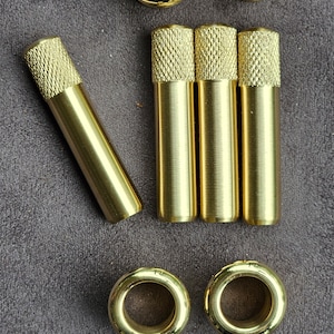 Puede incluir: Un juego de ojales y arandelas de metal de color dorado. Los ojales son redondos con un agujero en el centro. Las arandelas son cil&iacute;ndricas con una parte superior moleteada.