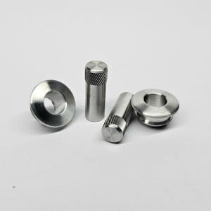 Puede incluir: Cuatro pequeños componentes metálicos plateados están dispuestos sobre una superficie blanca. Dos son cilíndricos con tapas texturizadas y dos son circulares con aberturas centrales. Piezas de máquina.